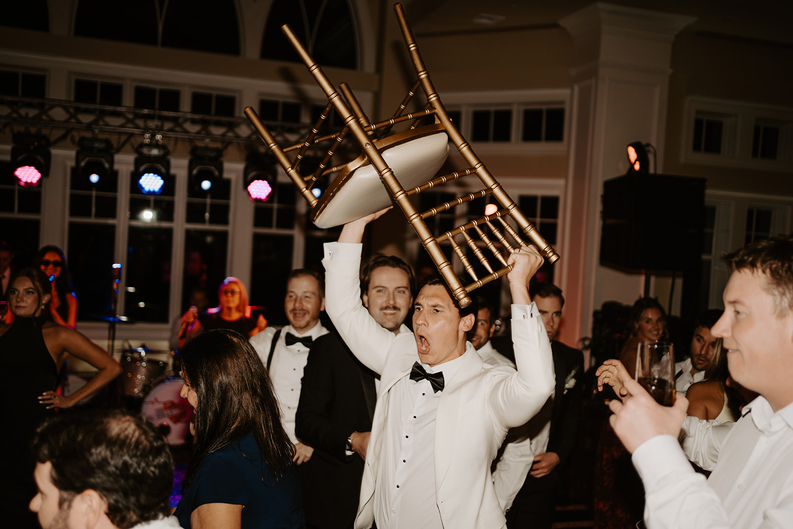 Groom in a white tux holding a gold chair high above his head in an energetic dance floor celebration.